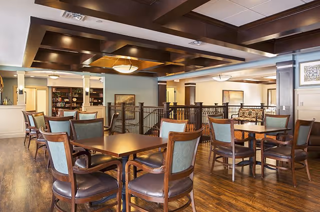 A spacious and well-lit common area with wooden flooring and ceiling beams. Several wooden tables with cushioned chairs are arranged throughout the room. The area includes a railing overlooking a lower level, and there are paintings and decorative elements on the walls. The overall ambiance is warm and inviting.