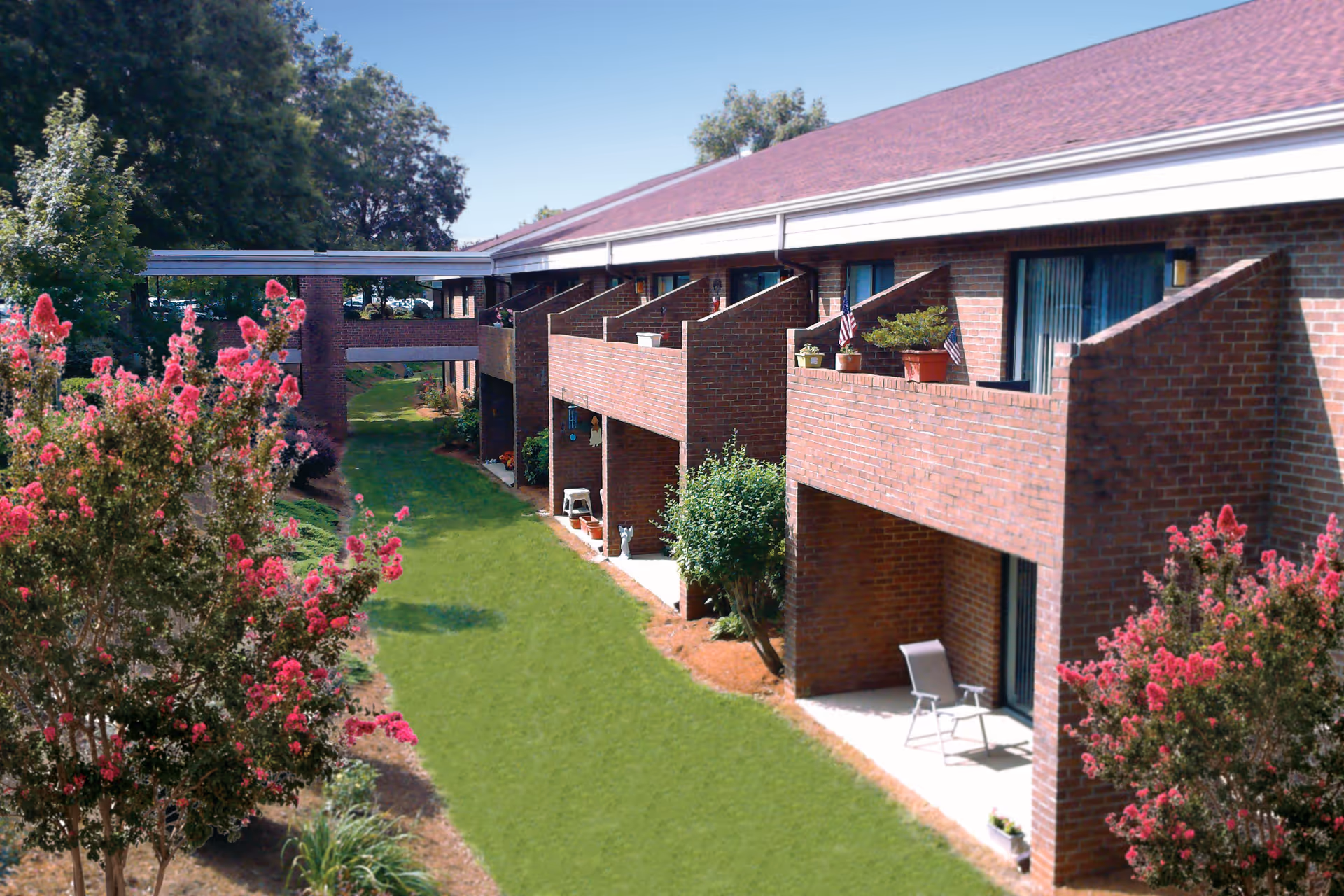 Brick two-story senior living building with balconies overlooking a grassy courtyard and blooming pink crepe myrtle trees.