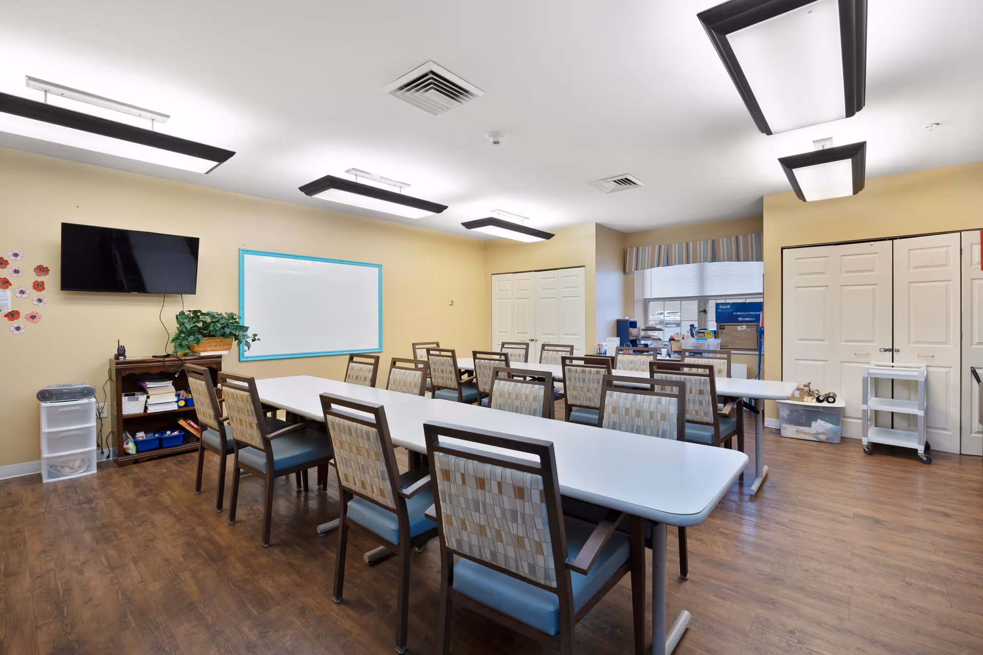 A bright multipurpose dining/activity room with long tables and chairs, a wall-mounted TV, whiteboard, and storage cabinets.