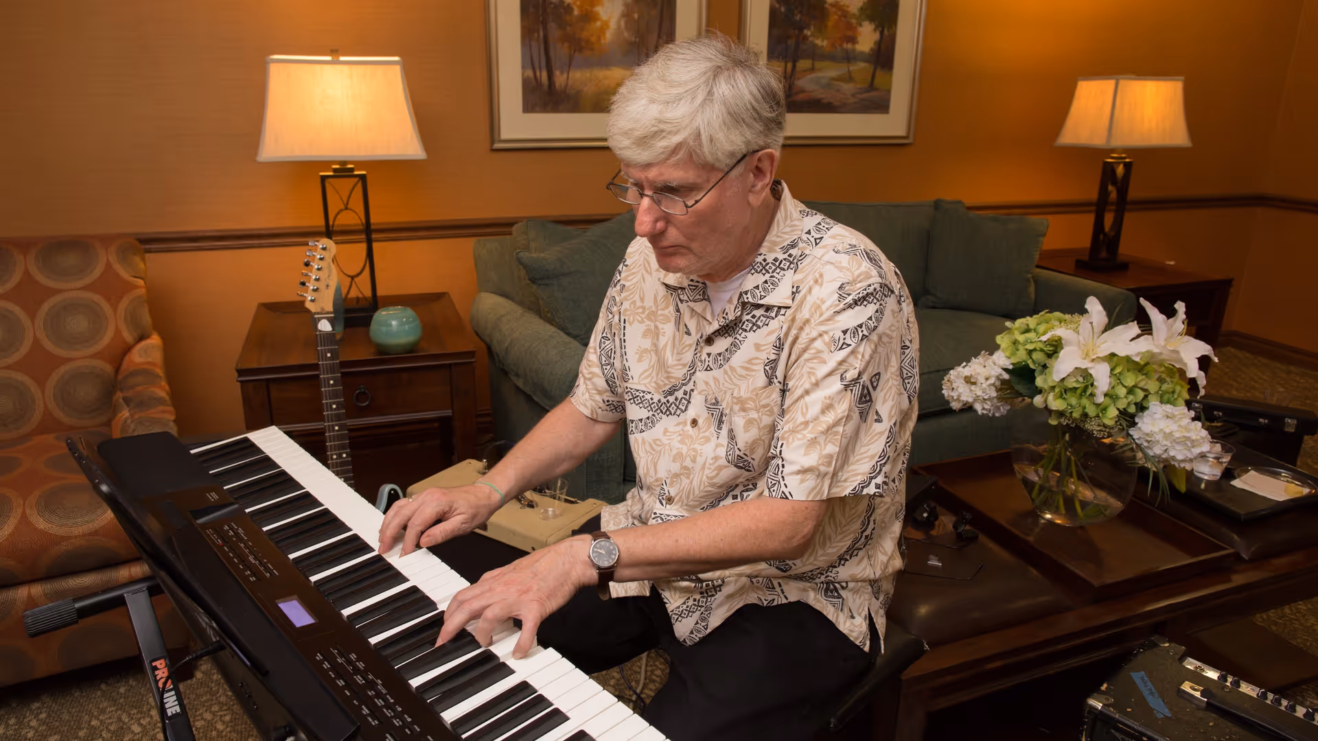 An elderly man with gray hair and glasses playing a keyboard piano in a warmly lit living room. The room features a green sofa, a wooden coffee table with a vase of white and green flowers, a side table with a lamp and a guitar resting against it, and a framed landscape painting on the wall.