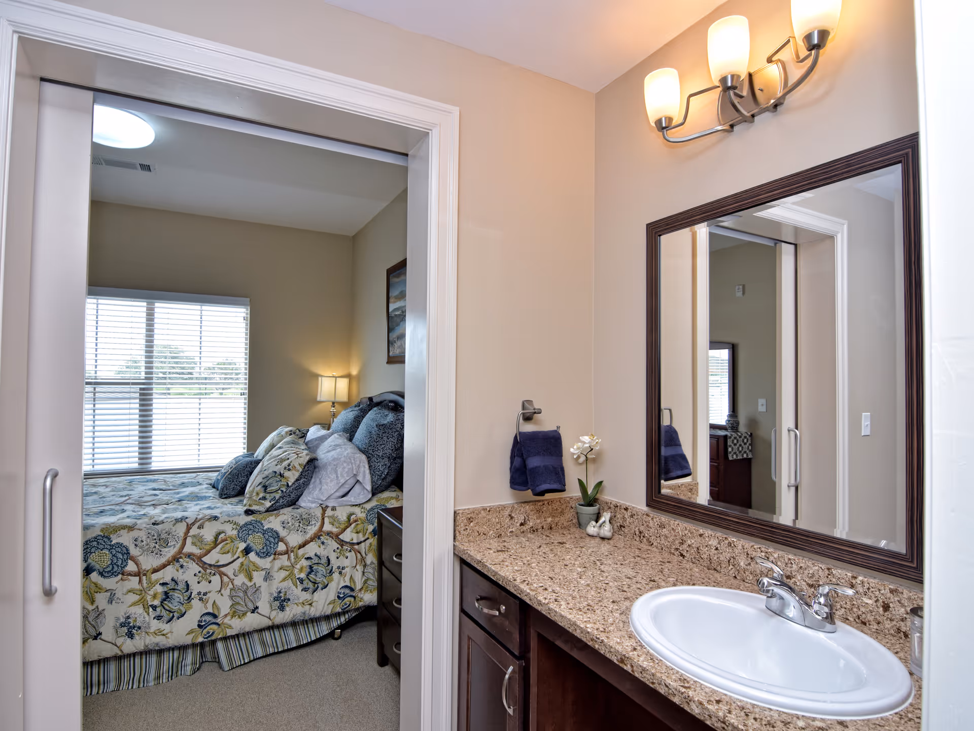 View of a bathroom vanity with a granite countertop, white sink, and a large mirror above it. A small potted plant and two decorative birds are on the countertop. A blue hand towel hangs on a ring next to the mirror. Through an open doorway, a bedroom is visible with a bed covered in a floral patterned bedspread and multiple pillows, a nightstand with a lamp, and a window with blinds letting in natural light.