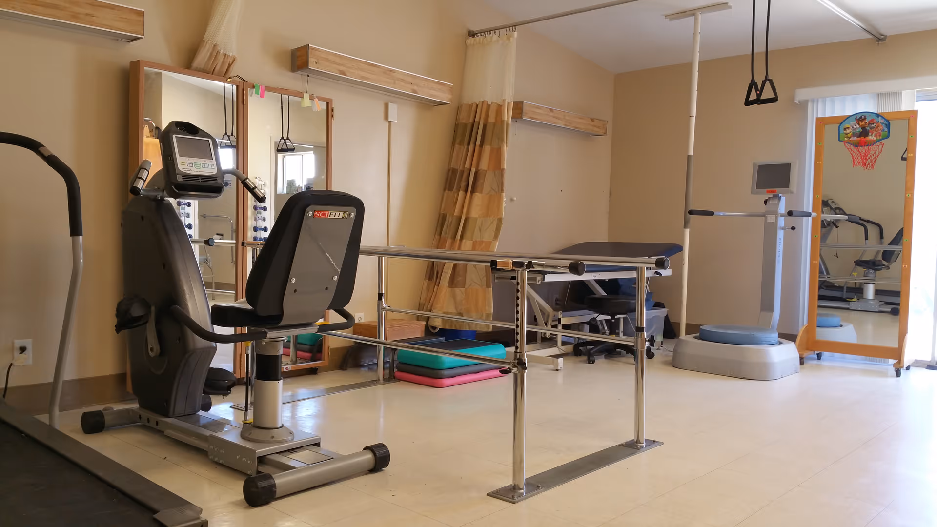 A physical therapy or exercise room with equipment including a recumbent exercise bike, parallel bars for walking support, a padded examination table, a balance platform, and a full-length mirror with a small basketball hoop attached. The room has beige walls, a curtain divider, and a tiled floor.