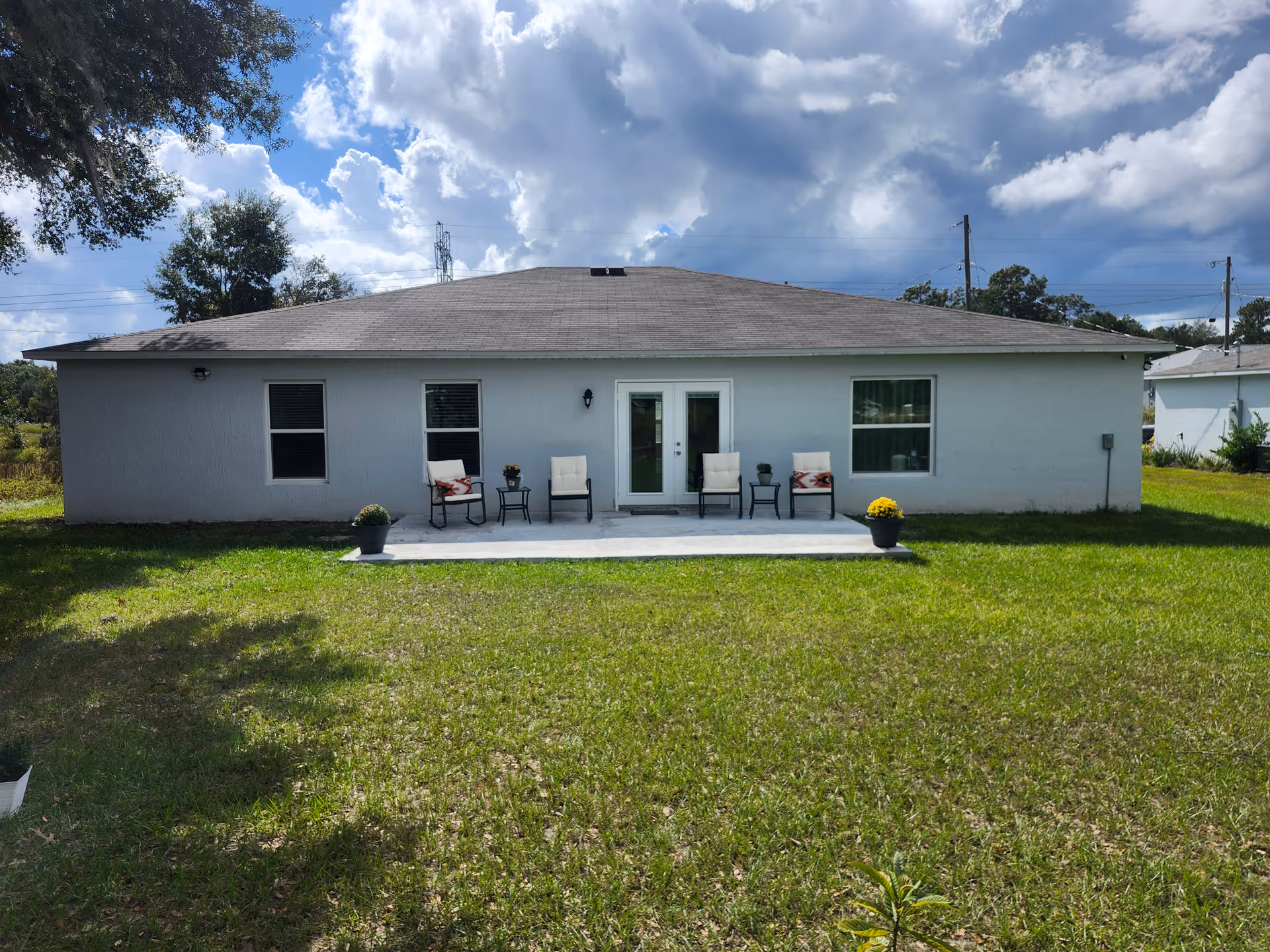 Back view of a single-story building with a gray roof and white walls. There is a small concrete patio with four cushioned chairs and two small tables, each table holding a potted plant. Two larger potted plants with yellow flowers are placed at the edges of the patio. The building has three windows and a set of double glass doors. The foreground shows a grassy lawn under a partly cloudy sky.