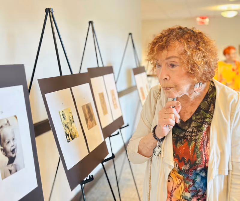 An elderly woman with curly hair and colorful clothing thoughtfully looks at a series of black and white photographs displayed on easels along a hallway wall inside a senior living facility.