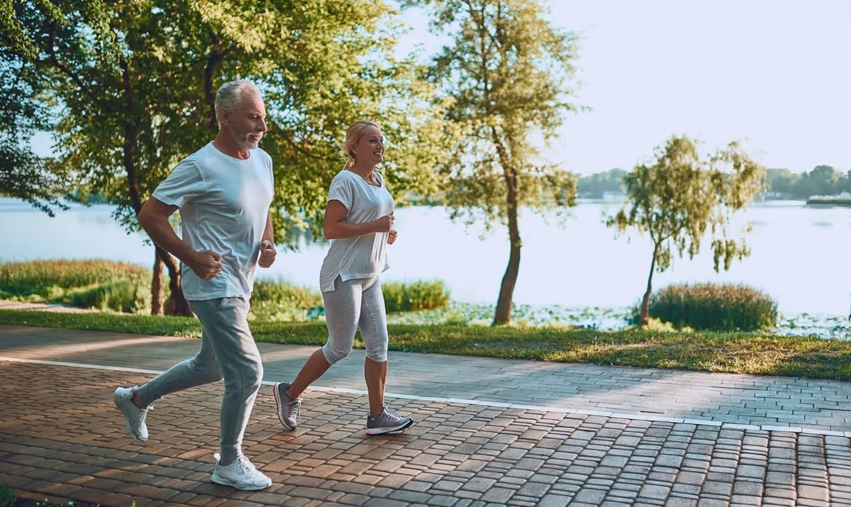 An older man and woman jogging together on a paved path near a lake with trees and greenery in the background on a sunny day.