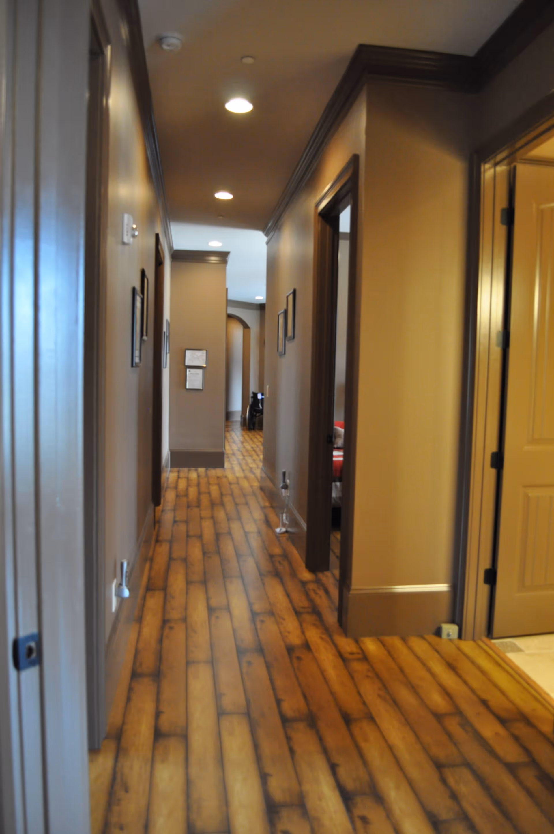 A long, narrow hallway with wooden plank flooring and beige walls. Several doorways line the hallway, some open, revealing parts of rooms. The ceiling has recessed lighting, and there are framed pictures hanging on the walls.