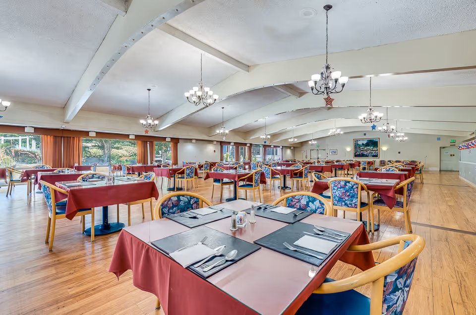 Spacious dining hall with rows of tables covered in red tablecloths, floral-upholstered chairs, chandeliers, and large windows.