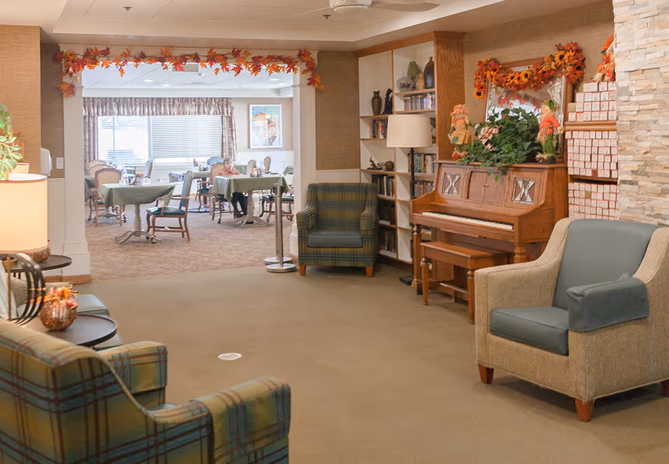 A cozy assisted living common area with comfortable armchairs, a wooden piano decorated with plants and autumn-themed garlands, and a bookshelf. In the background, there is a dining room with tables covered in green tablecloths and people seated, enjoying their time.