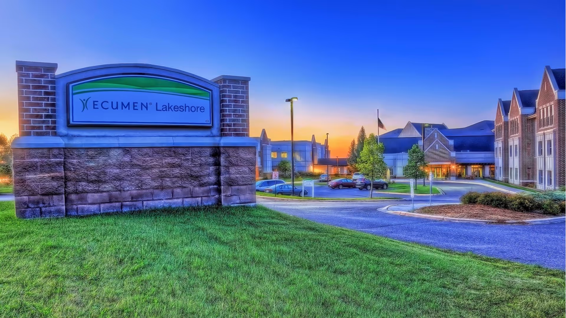 Exterior view of Ecumen Lakeshore facility at sunset with a large stone sign in the foreground and the building, parking lot, and trees in the background.