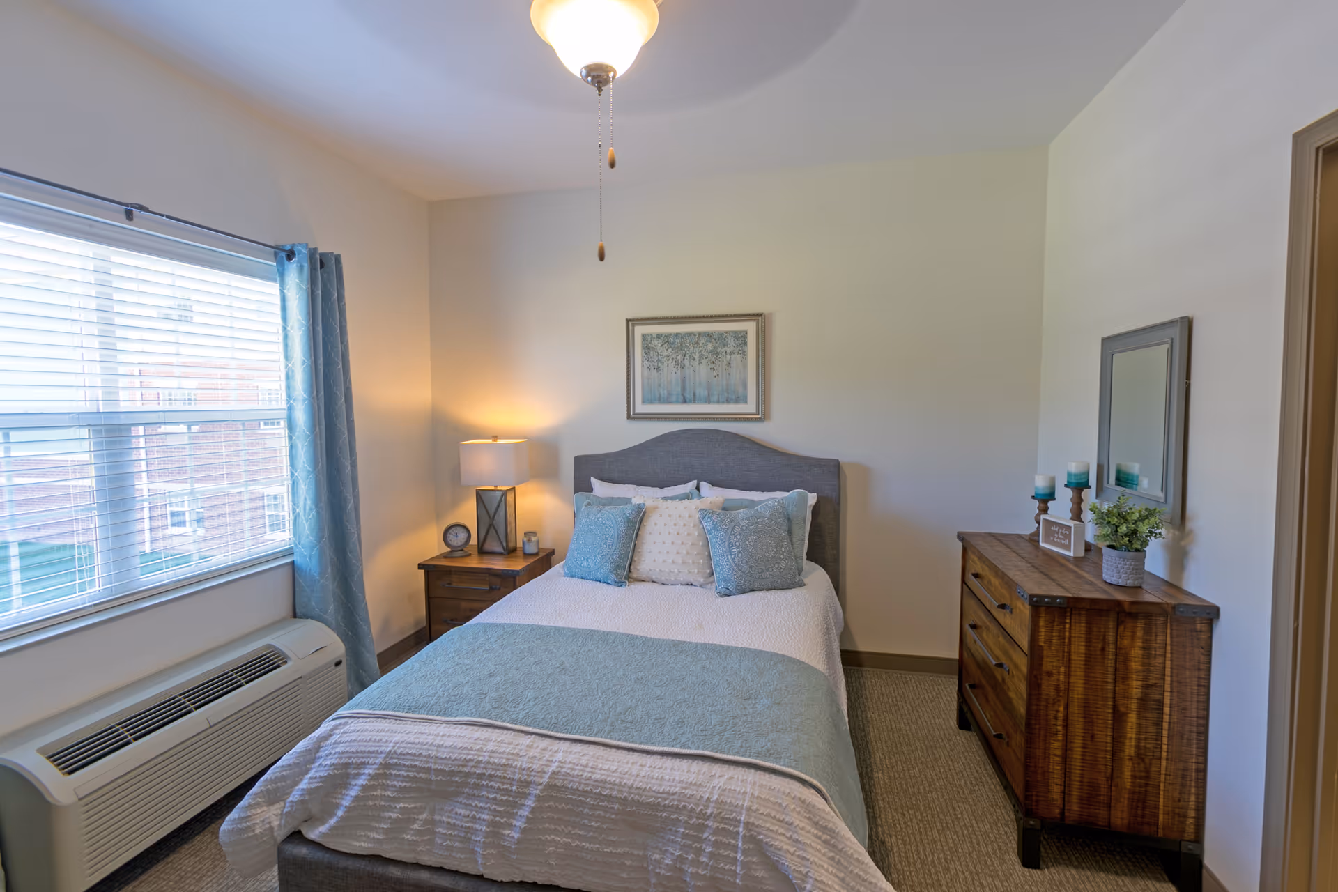 A cozy bedroom with a neatly made bed featuring white and light blue bedding and pillows. There is a wooden nightstand with a lamp and clock on the left side of the bed, and a wooden dresser with a mirror, candles, and a small plant on the right side. A large window with blue curtains allows natural light to fill the room.