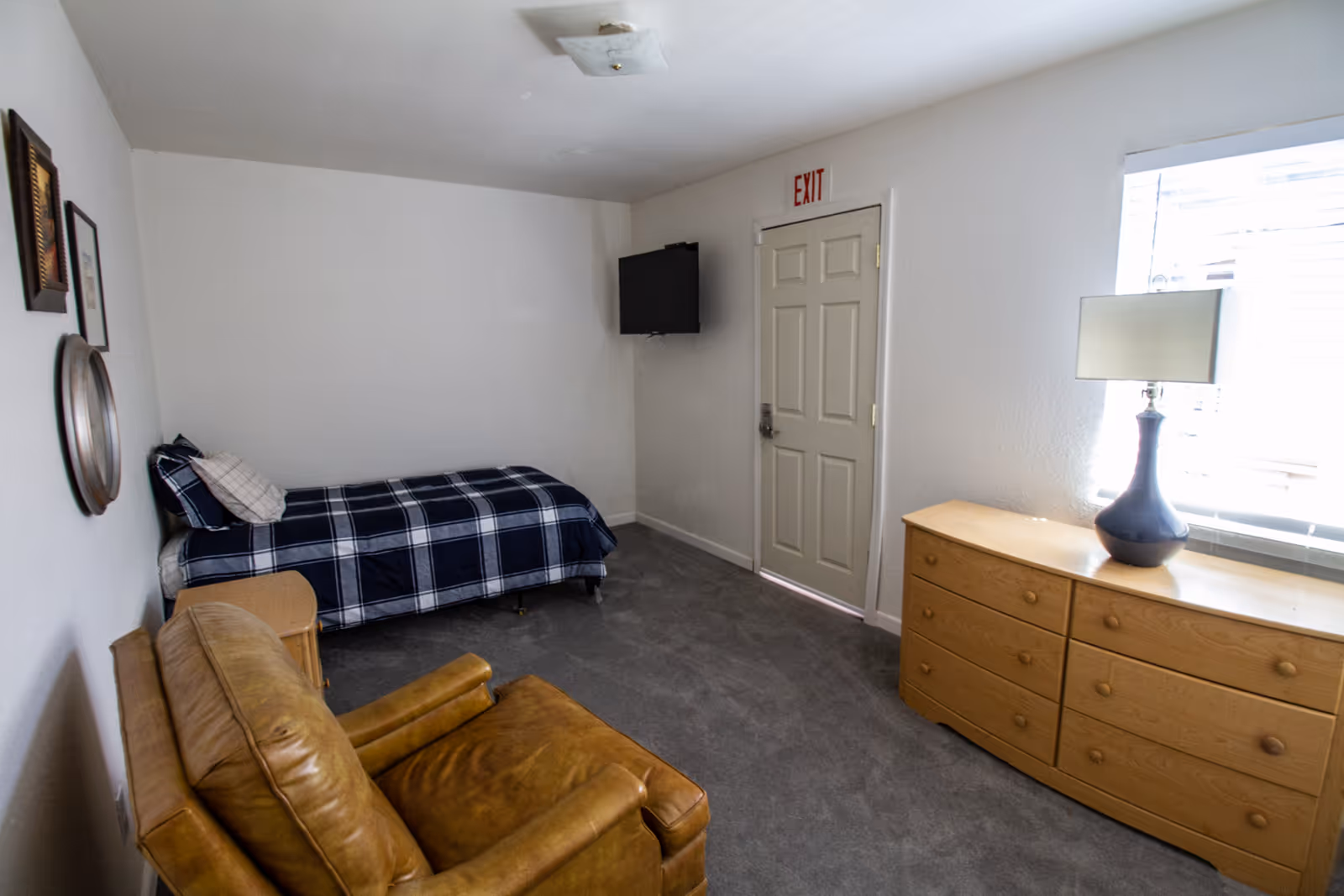 Small furnished bedroom with a single bed, leather armchair, dresser with lamp, wall-mounted TV, and an exit door.