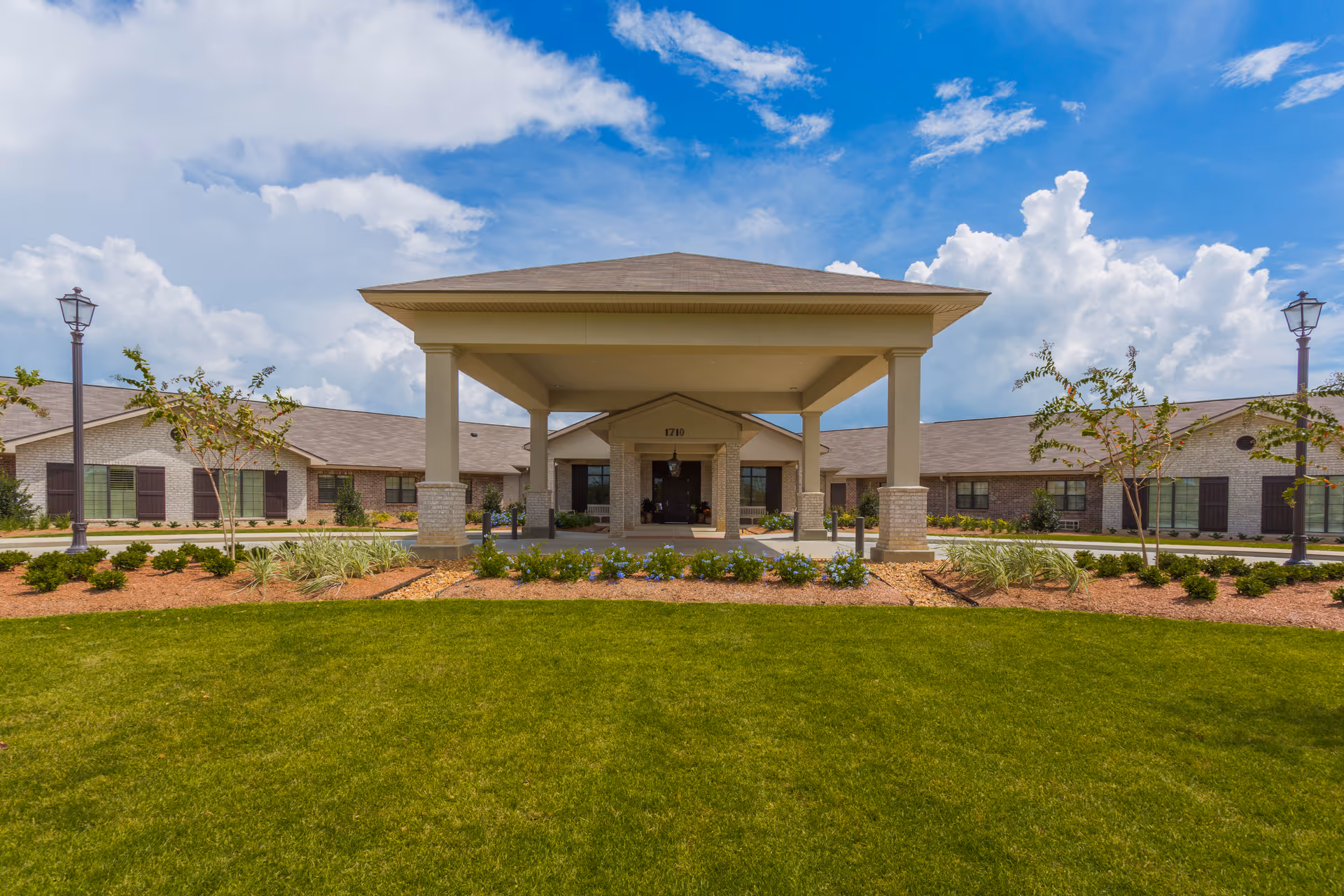 Front exterior view of Landmark Of Acadiana facility with a covered entrance, manicured lawn, small trees, and lampposts under a partly cloudy blue sky.