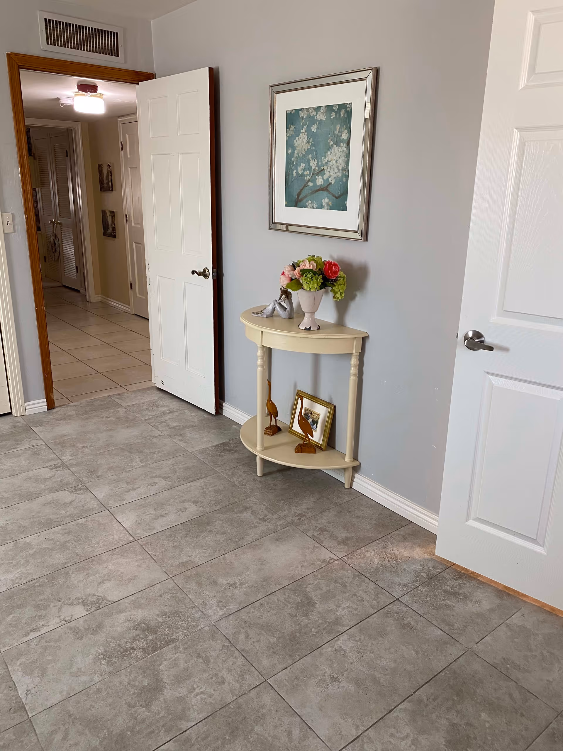 A hallway area with gray tiled flooring and light gray walls. There is a small cream-colored half-moon table against the wall with a vase of flowers, two wooden bird figurines, and a framed photo on the lower shelf. Above the table hangs a framed artwork featuring white blossoms on a blue background. Two white doors are partially open, one leading to a hallway with beige tiled flooring and another room. The hallway has a ceiling light fixture and some wall decorations.