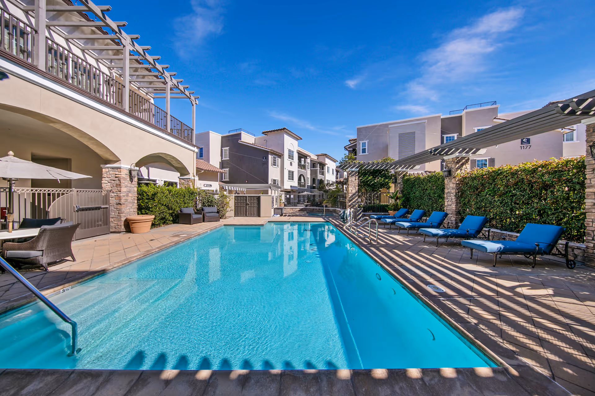 Outdoor swimming pool area at Meridian at Lake San Marcos with clear blue water, surrounded by lounge chairs with blue cushions, shaded pergolas, and residential buildings in the background under a bright blue sky.
