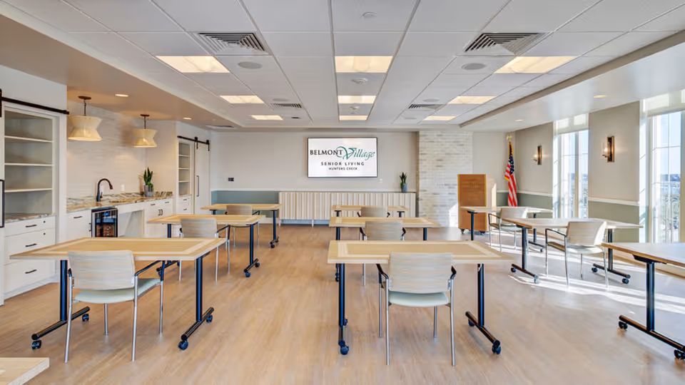 A bright and spacious room with multiple tables and chairs arranged for individual seating. The room features large windows allowing natural light to fill the space, a kitchenette area with cabinets, a sink, and a small refrigerator on the left side. A screen on the far wall displays the Belmont Village Senior Living Hunters Creek logo. An American flag stands in the corner near the windows.