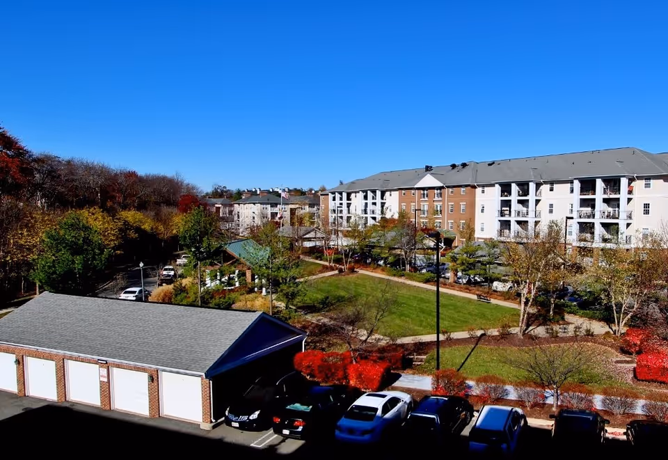 View of an outdoor area at Kentlands Manor featuring a large green lawn with walking paths, surrounded by trees and bushes with autumn foliage. In the background, there is a multi-story residential building with balconies. In the foreground, there is a row of garages and parked cars under a clear blue sky.