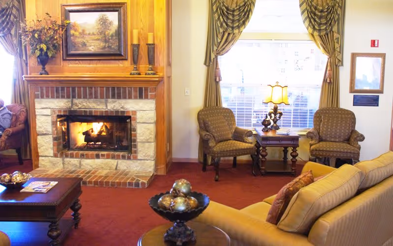 A cozy living room with a lit stone fireplace, upholstered armchairs and sofa, coffee tables, and a window with draped curtains.