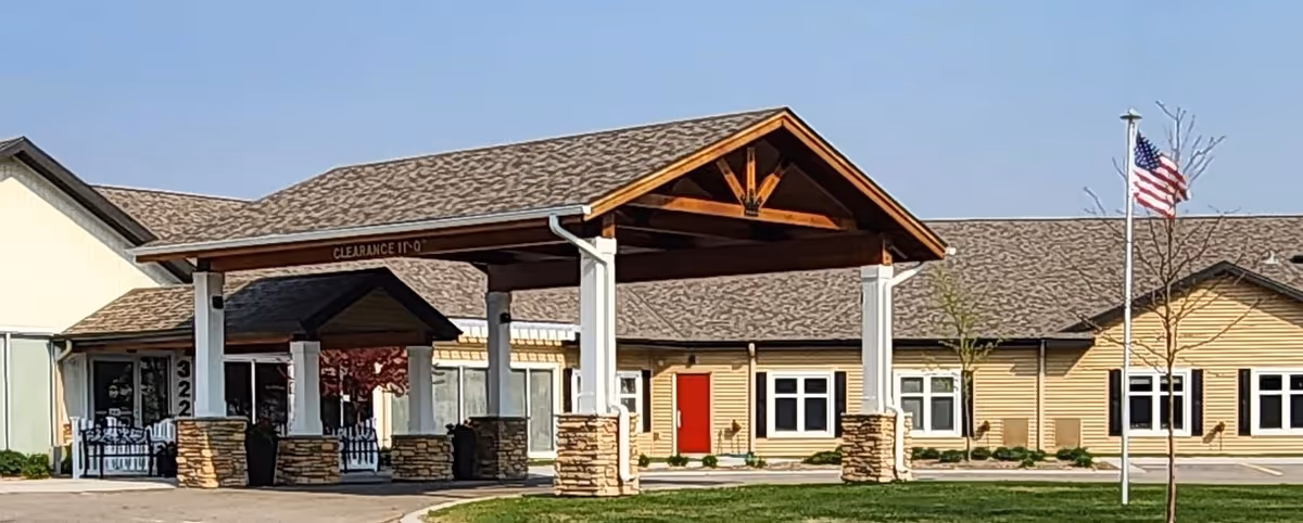 Front exterior view of a single-story senior living facility building with a covered entrance supported by white columns with stone bases. The building has beige siding, multiple windows, a red door, and an American flag on a flagpole in the front lawn.