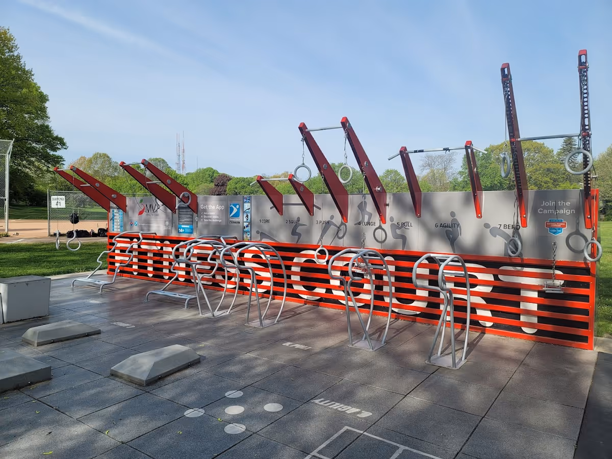 Outdoor fitness court with various exercise stations including pull-up bars, gymnastic rings, and workout benches. The court has a red and black striped wall with the words 'FITNESS COURT' prominently displayed. There are instructions for different exercises on the wall and a baseball field visible in the background.