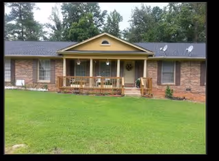 Single-story brick house with a front porch featuring wooden railings and steps, surrounded by a well-maintained green lawn and tall trees in the background.