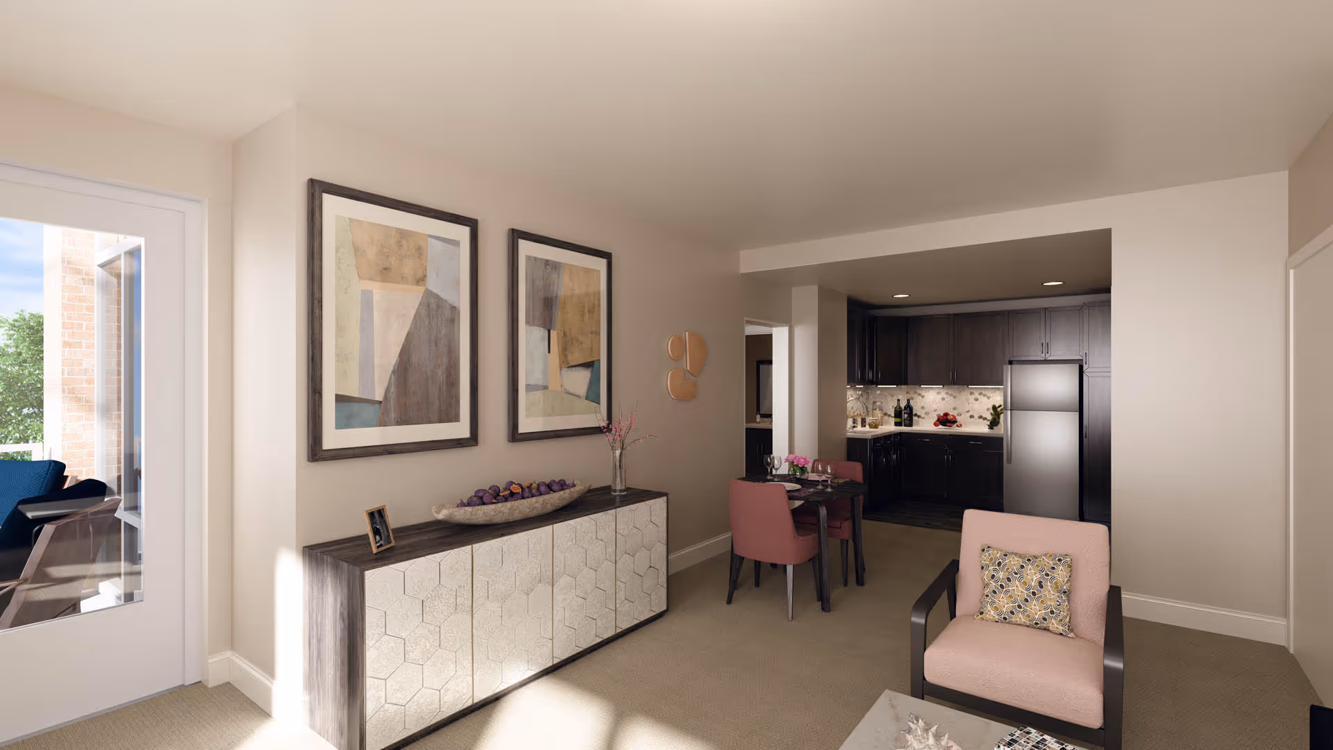 Interior view of a senior living facility showing a living room area with a pink armchair and a small table, a dining area with a table set for two and pink chairs, and a kitchen with dark cabinets and a stainless steel refrigerator. There are two abstract paintings on the wall above a sideboard, and a glass door leading to an outdoor patio with seating.
