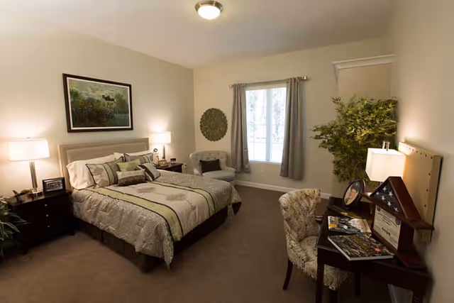 A cozy bedroom with a neatly made bed featuring patterned bedding and multiple pillows. There are two bedside tables with lamps on either side of the bed. A framed picture hangs above the bed. A cushioned chair is placed near a window with gray curtains. A desk with a lamp, framed certificate, folded American flag in a display case, and magazines is positioned against the wall, accompanied by a patterned chair. A large green plant is in the corner of the room.