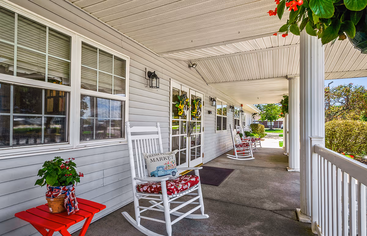 Covered front porch with white rocking chairs, potted plants, wreaths, and columns leading to the building entrance.