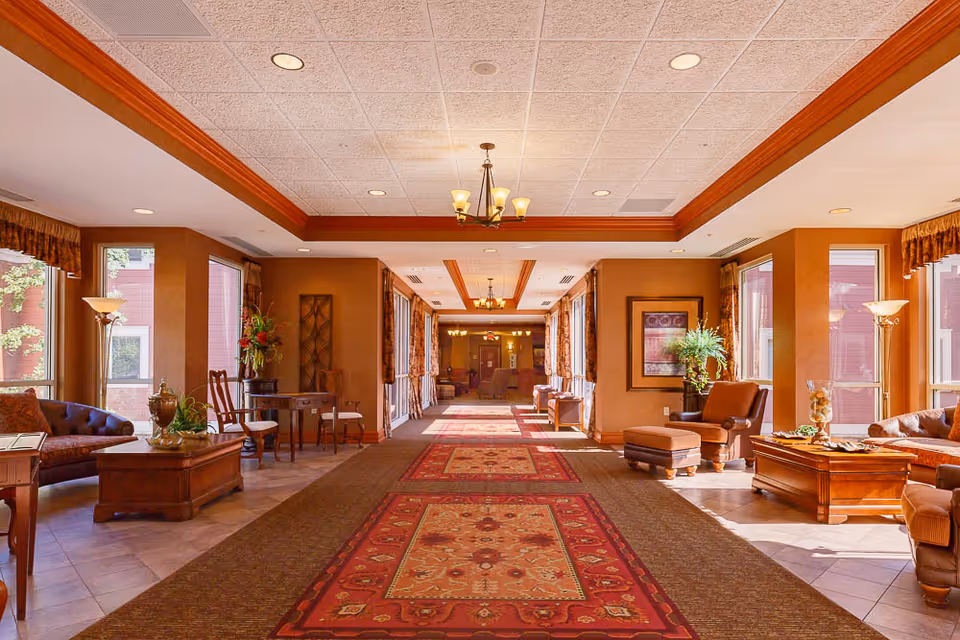 A spacious and well-lit interior hallway of a senior living facility with large windows on both sides, allowing natural light to fill the space. The hallway features a long red patterned carpet runner, wooden furniture including chairs, tables, and sofas arranged along the sides, decorative plants, framed artwork on the walls, and ceiling lights with chandeliers.