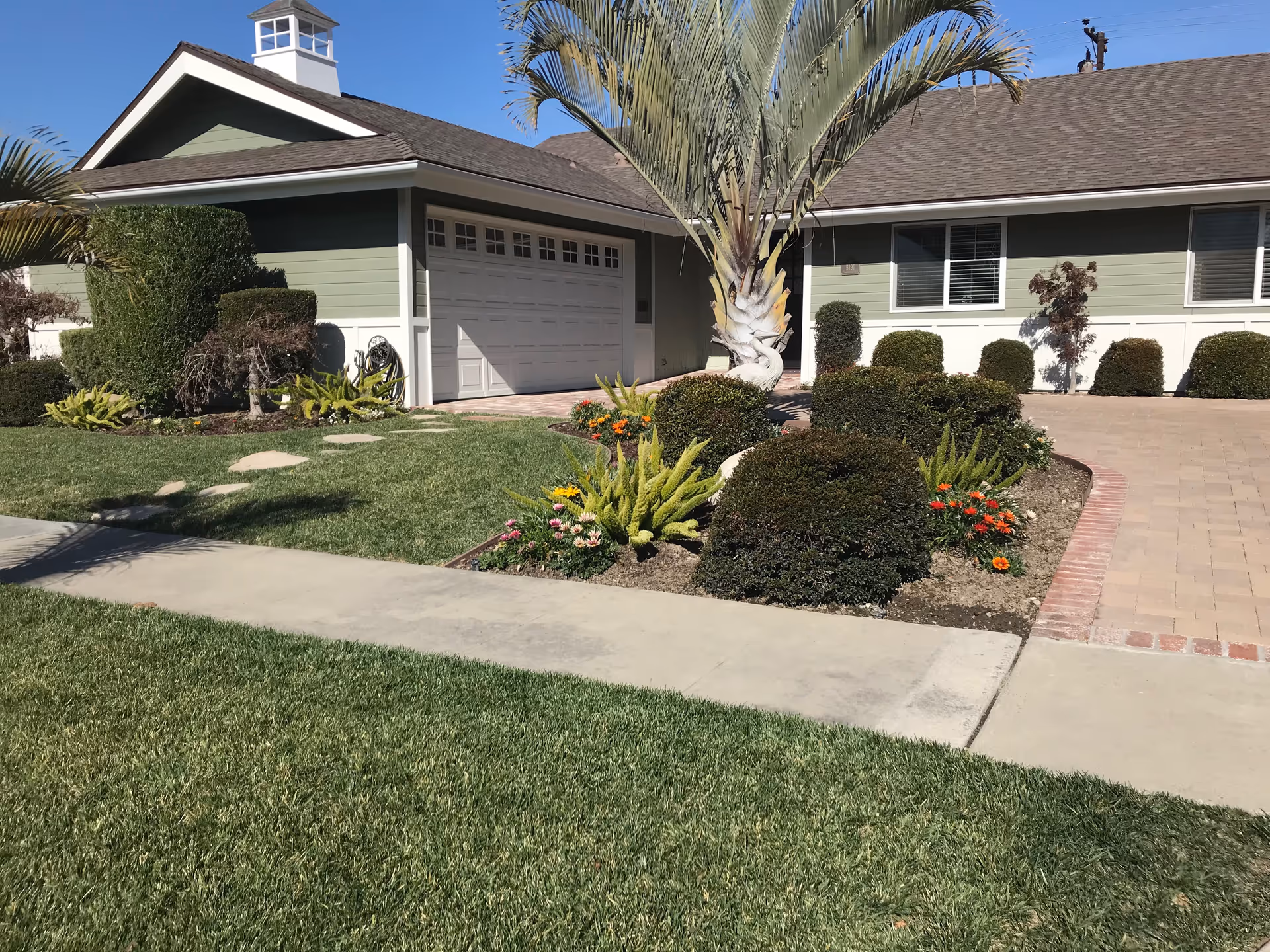 Front exterior of a single-story house with a garage, driveway, palm tree, manicured lawn, and flower beds.