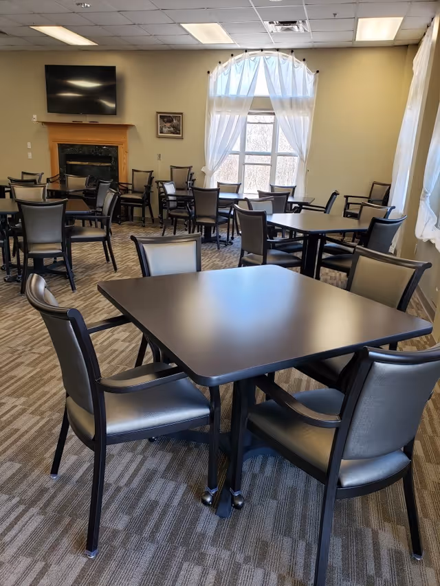 A dining room with multiple square tables and chairs arranged neatly. The room has beige walls, a carpeted floor, a large window with white curtains letting in natural light, a wall-mounted TV above a wooden fireplace, and a framed picture on the wall.
