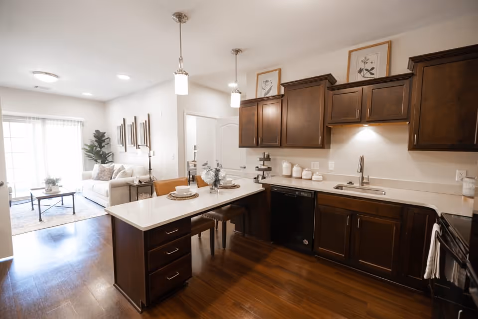 Open-concept kitchen with dark wood cabinets, a white island with pendant lights and seating, and a living area with a sofa and coffee table in the background.