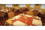 Dining room with multiple tables set for meals, upholstered chairs, yellow walls, and patterned carpet.