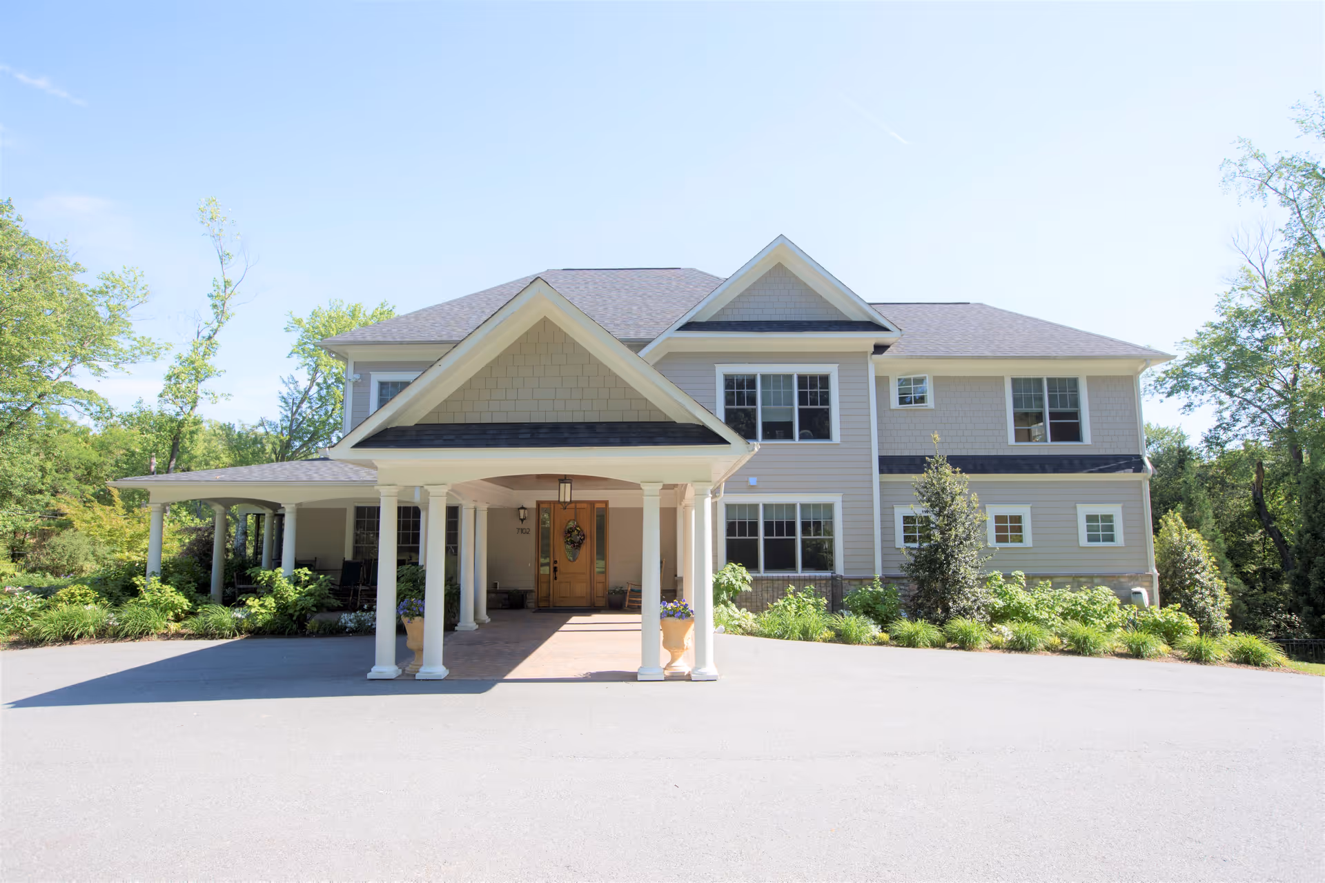 Front exterior view of a two-story residential building with a covered entrance supported by white columns, surrounded by greenery and trees under a clear blue sky.