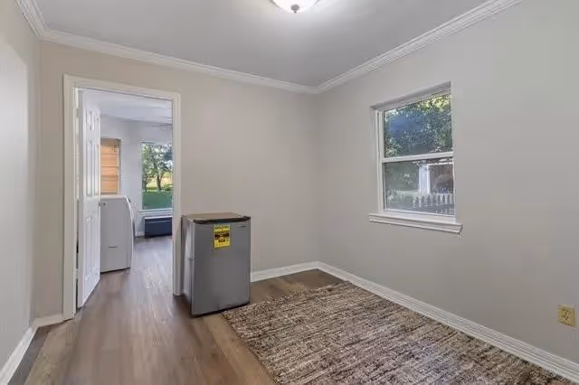 An empty room with light gray walls, a window showing greenery outside, a small refrigerator, and a brown patterned rug on wooden flooring. An open door leads to another room with a washing machine and a view of the outdoors.