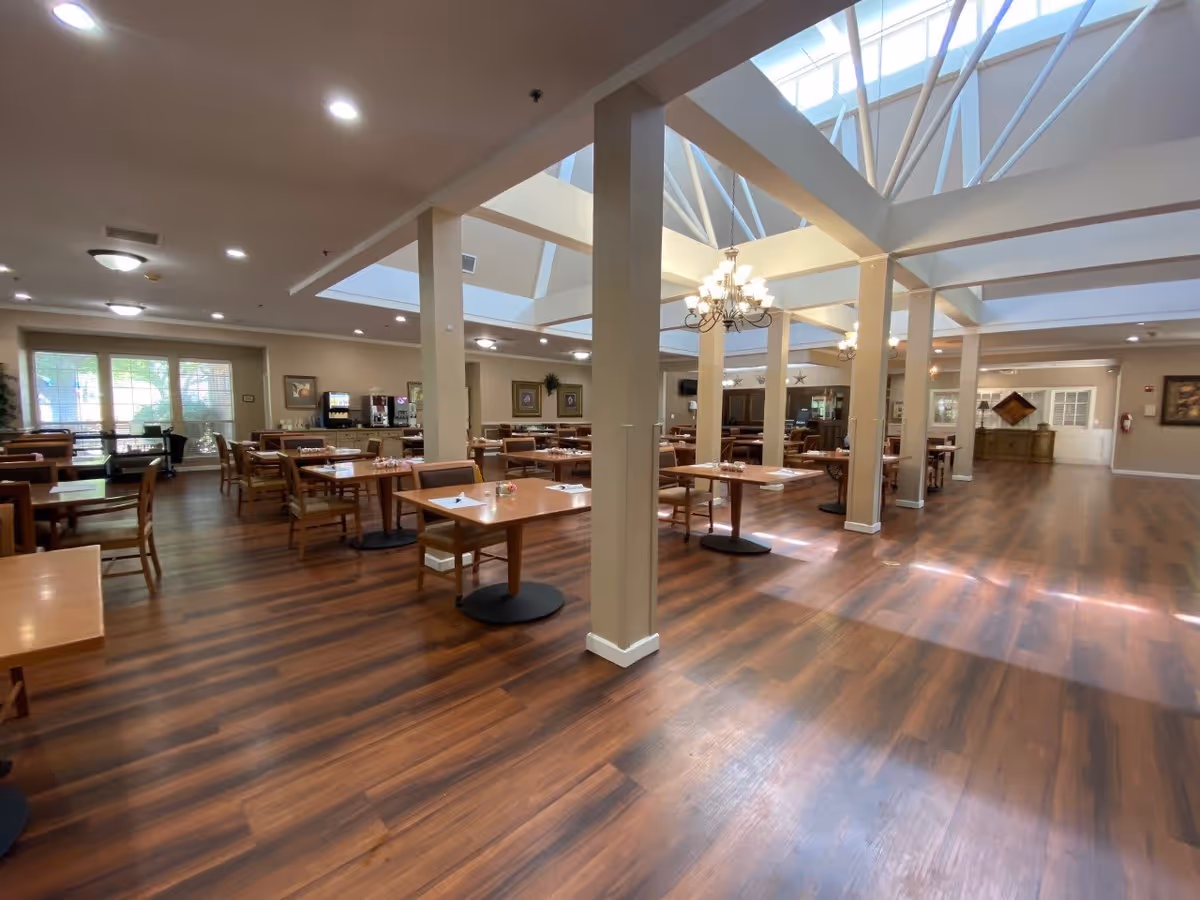 Spacious dining room with wooden floors, multiple tables and chairs arranged neatly, large windows letting in natural light, and ceiling with skylights and chandeliers.