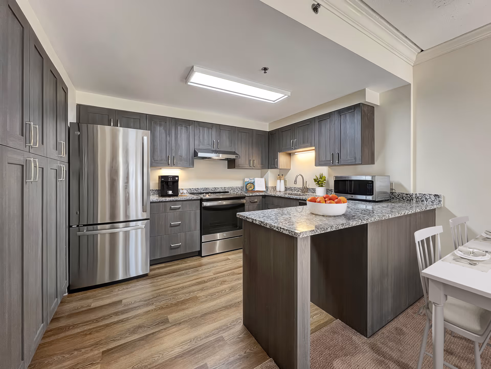 Modern kitchen with gray wooden cabinets, stainless steel refrigerator and oven, granite countertops, a coffee maker, a microwave, and a bowl of apples on the island. The floor is wood, and there is a small dining table with white chairs partially visible on the right.