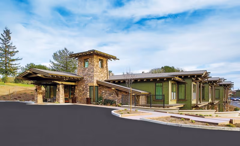 Exterior view of a single-story building with green siding and stone accents, featuring a covered entrance supported by stone pillars, surrounded by a paved driveway and landscaped areas with small trees and shrubs under a partly cloudy sky.