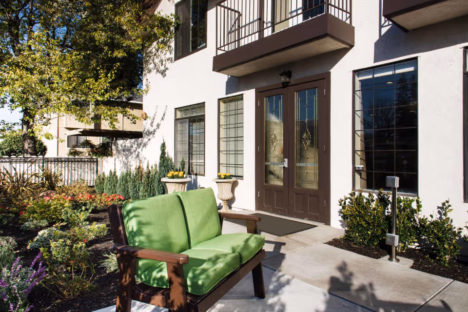 Outdoor seating area in front of a building with white walls and large windows. There is a green cushioned wooden bench on a concrete pathway surrounded by landscaped plants and flowers. The building has double glass doors with decorative glass panels and small balconies above.