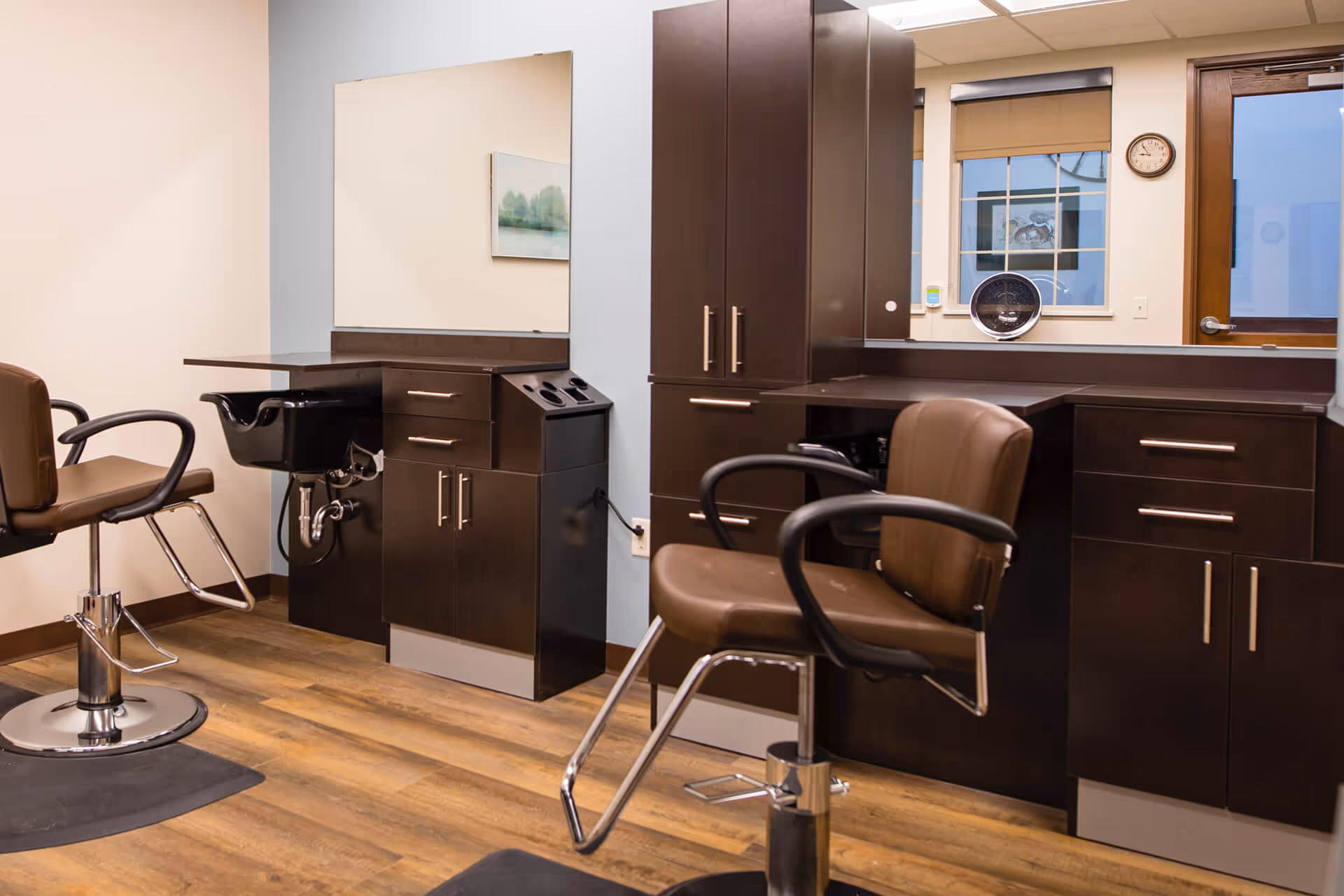 Interior of a salon area with two brown salon chairs, dark wood cabinetry, a black hair washing sink, a large mirror, and a window with a view outside. The floor is wooden and the walls are light-colored.