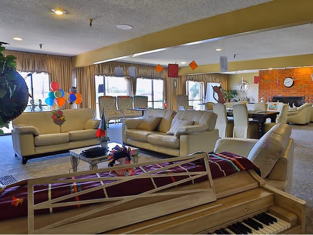 Bright communal lounge with sofas, armchairs, a dining table, and a piano in the foreground near large windows and balloons.