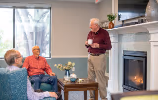 Three elderly men in a cozy living room setting. Two men are seated on chairs near a window with natural light, while the third man stands holding a cup near a fireplace with a TV mounted above it. The room has a coffee table with decorative items and a plant on the mantel.