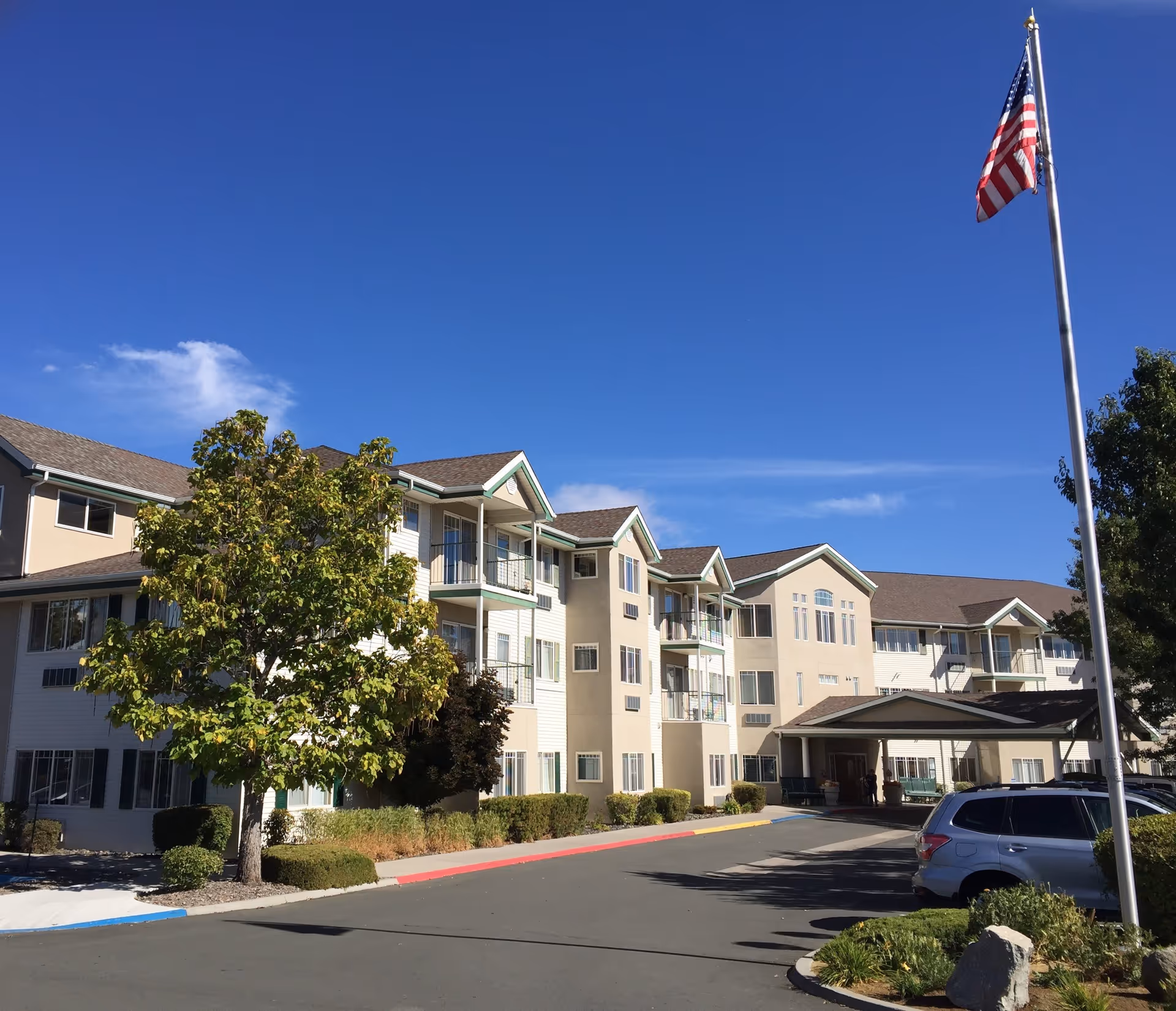 Exterior view of a multi-story retirement community building under a clear blue sky, with a large tree and an American flag on a flagpole in the foreground. Several cars are parked near the entrance.
