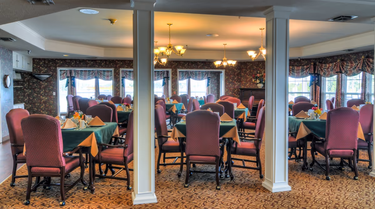 A dining room with multiple tables covered in green and beige tablecloths, each set with napkins, cups, and small flower arrangements. The room has patterned carpet, floral wallpaper, large windows with floral curtains, and several chandeliers providing warm lighting.