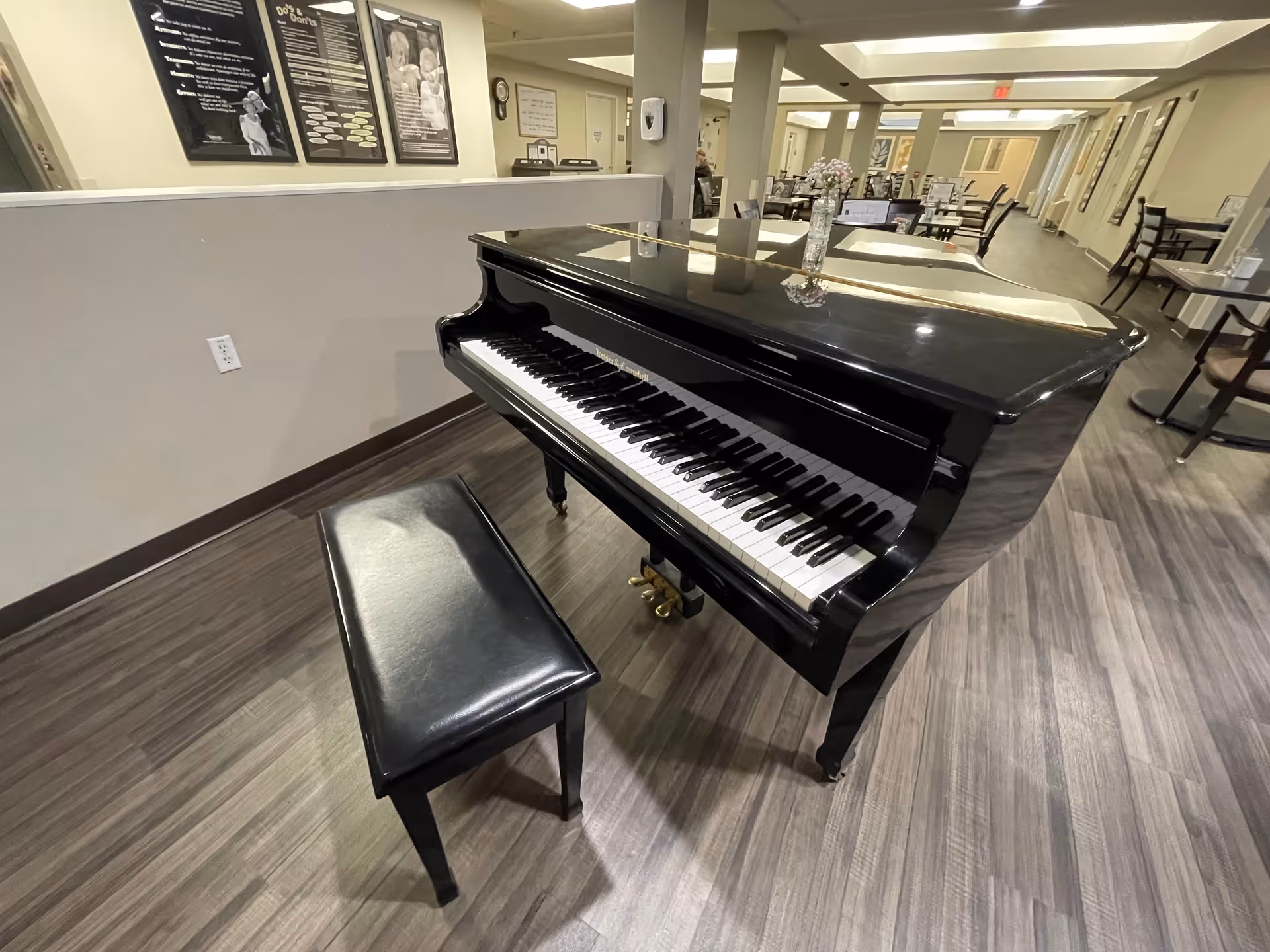 A black grand piano with a matching black bench is placed on a wood-patterned floor in a spacious room. The room has tables and chairs arranged in the background, with some wall decorations and informational posters visible. The lighting is bright and the overall setting appears to be a common area in a senior living facility.
