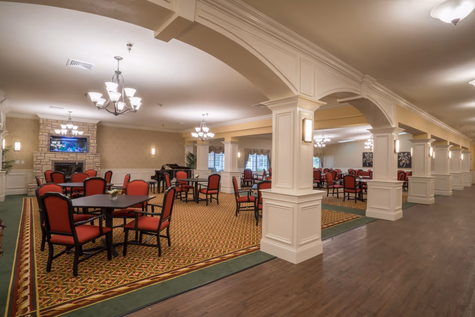 Spacious dining room with multiple tables and red cushioned chairs arranged on patterned carpet. The room features white decorative columns and arches, chandeliers hanging from the ceiling, a stone fireplace with a built-in aquarium above it, and a grand piano in the corner. Large windows with drapes allow natural light into the room.