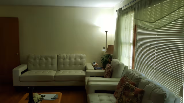 A living room with three white cushioned sofas arranged around a wooden coffee table. There is a floor lamp and a table lamp near the corner, a side table with some items on it, and large windows with blinds partially closed letting in natural light.