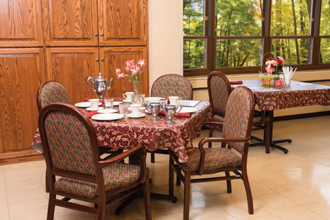 A dining area with two tables covered in patterned red tablecloths. Each table is set with white cups, saucers, plates, and silverware. One table has a silver coffee pot and a vase with pink flowers. The chairs around the tables have wooden frames and patterned upholstery. Large windows in the background show green trees outside, and wooden cabinets are visible along one wall.