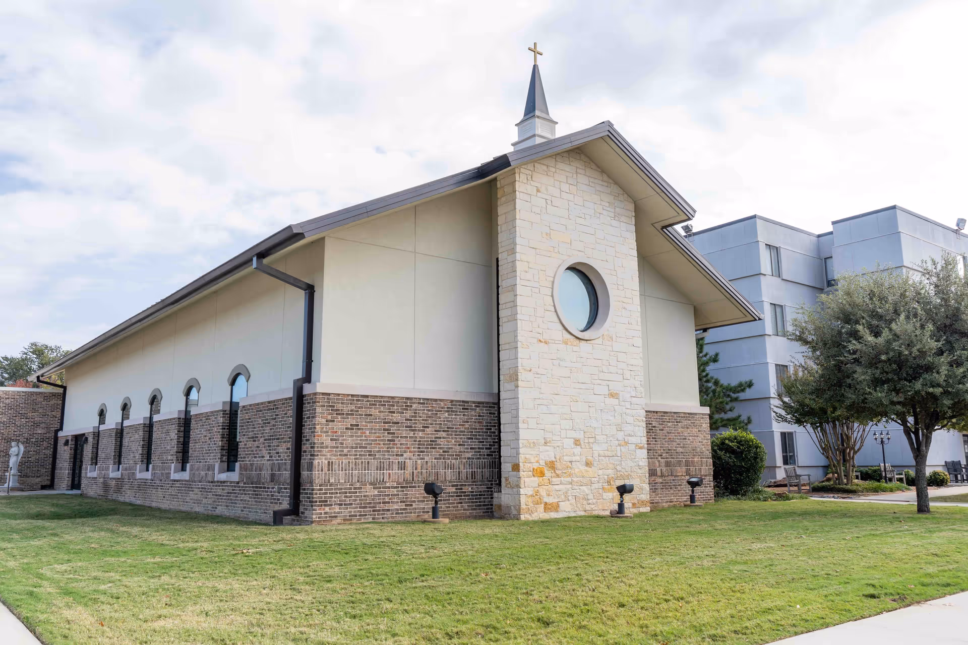 Exterior view of a building with a stone and brick facade, featuring a small steeple with a cross on top. The building is surrounded by a well-maintained lawn and some trees, with another multi-story building visible in the background.