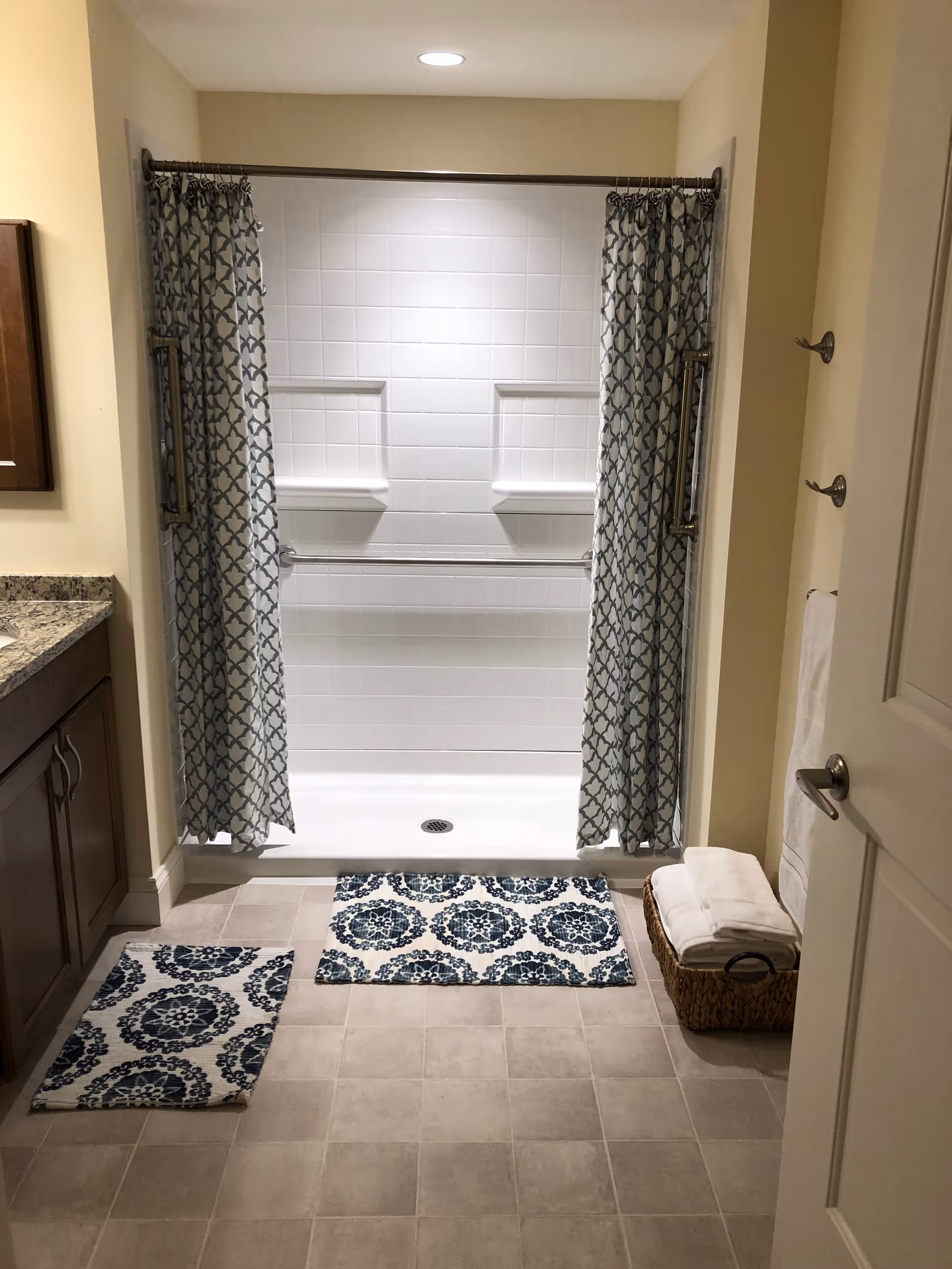 A clean bathroom with a tiled shower area featuring two built-in shelves and a shower curtain with a geometric pattern. The floor has two matching blue and white patterned rugs. To the right, there is a basket with folded white towels and a towel hanging on a hook on the wall. On the left side, there is a granite countertop with wooden cabinets underneath.