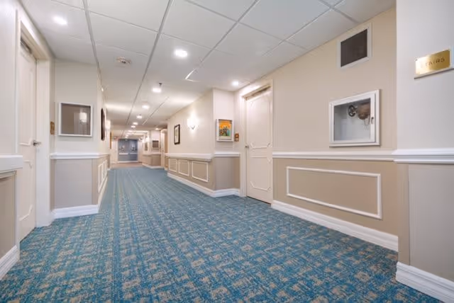 A well-lit indoor hallway with beige walls and blue patterned carpet. The hallway has several closed doors on both sides, framed artwork on the walls, and ceiling lights. There is a sign on the right wall that reads 'STAIRS'.